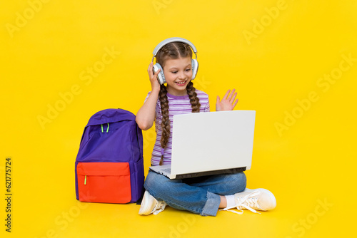 A schoolgirl sits with a laptop and communicates online with a teacher and a tutor. Distance education. A young girl is sitting cross-legged on the floor. Yellow isolated background.