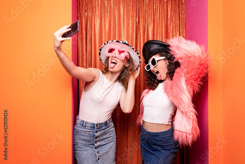 Attractive couple of casual caucasian women posing with carnival dress having fun standing with yellow - orange background hugging and making face..Two ladies making a selfie.