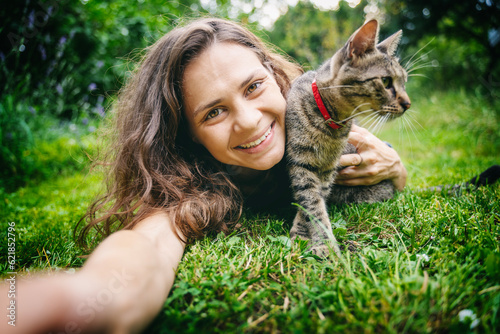 Happy young woman taking selfie with cute gray cat lying on green grass in summer garden