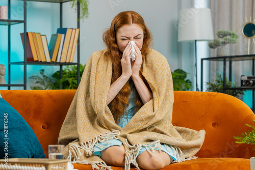 Young ill redhead woman suffering from cold or allergy sitting on sofa in living room at home. Sick girl wrapped in plaid blows sneezes wipes snot into paper napkin. Coronavirus quarantine, pandemic