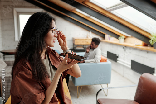 Young Asian businesswoman with mobile phone by her mouth recording voice message and looking through window in loft coworking space