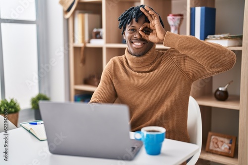 Young african man with dreadlocks working using computer laptop doing ok gesture with hand smiling, eye looking through fingers with happy face.
