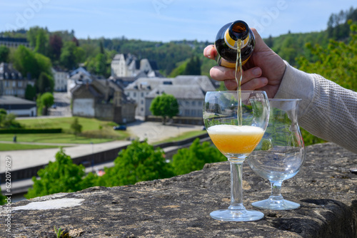 Pouring of blond strong Belgian abbey beer in to glass in sunny day with nice view on old town Bouillon, Belgium