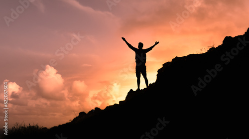 Hiker with arms raised on a mountain facing beautiful sunset silhouette against cloudy sky.