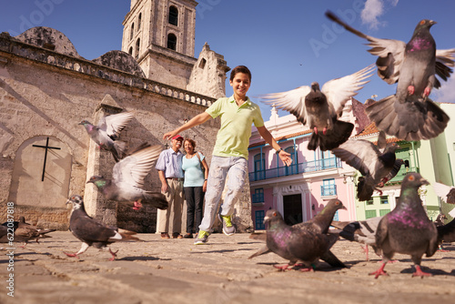 Happy tourist on holidays during vacation trip. Hispanic people traveling in Havana, Cuba. Grandpa and grandson feeding birds, with child running chasing pigeons