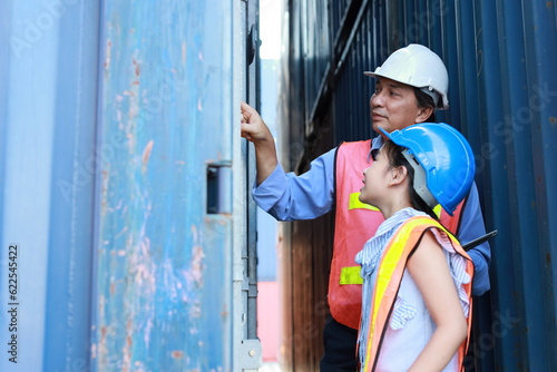 Asian foreman father and his daughter teaching little girl with tablet. Engineer dad with safety hat pointing at container and show something to his kid at industrial container cargo freight ship