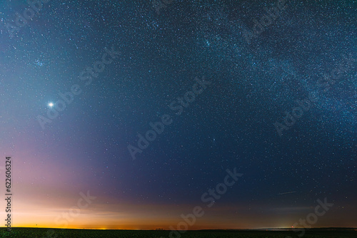 Night Starry Sky With Glowing Stars Above Countryside Field Landscape In Early Spring. Bright Glow Of Planet Venus In Sky Among The Stars. Sky In Warm Lights Of Evening Sunset Dawn Or Morning Sunrise.