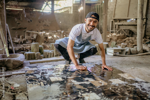 Smiling stonemason squats while washing large squares of stone in the workshop
