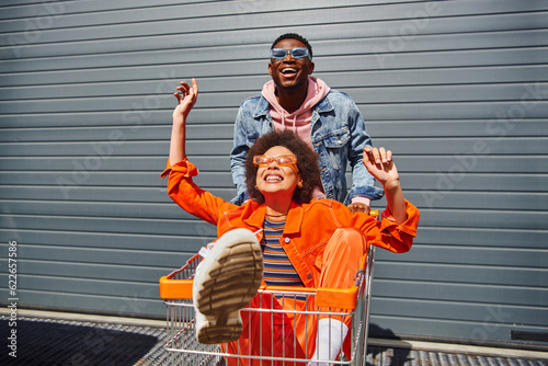 Cheerful young african american best friends in sunglasses and bright outfits having fun with shopping cart and spending time near building at background, friends hanging out together