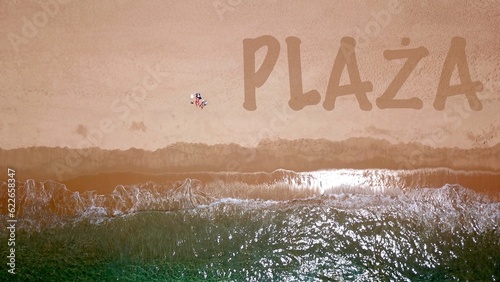 Widok z lotu ptaka na piękną piaszczystą plażę nad błękitnym morzem, gdzie leży i opala się dwóch wczasowiczów, obok nich napis "PLAŻA" na piasku