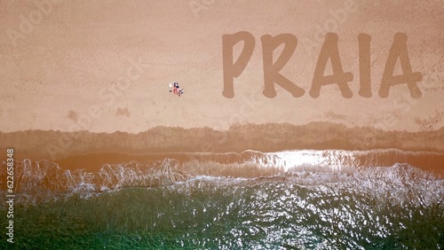 Vista aérea de uma bela praia de areia junto ao mar azul, onde dois veranistas estão deitados e tomando banho de sol, ao lado deles a inscrição "PRAIA" na areia