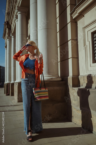 Fashionable woman wearing trendy orange linen shirt, crochet crop top, maxi denim skirt, blue sunglasses, holding wicker tote bag, posing in street of European city. Outdoor full body fashion portrait