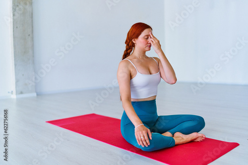 Сalm concentrated relaxed redhead woman with closed eyes practices yoga breathing exercise on mat in studio class