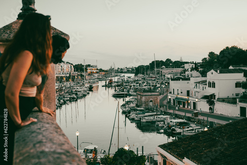 Widok na duży port z jachtami oraz patrzący na niego ludzie o zachodzie słońca na wyspie Menorca