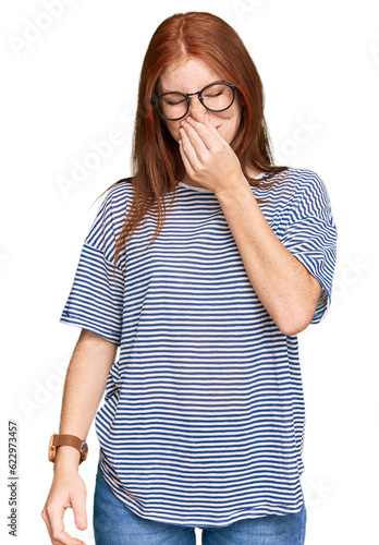 Young read head woman wearing casual clothes and glasses smelling something stinky and disgusting, intolerable smell, holding breath with fingers on nose. bad smell