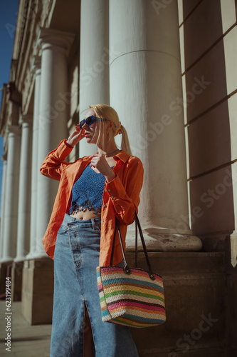 Fashionable woman wearing trendy orange linen shirt, crochet crop top, maxi denim skirt, blue sunglasses, holding striped wicker tote bag, posing in street of European city. Outdoor fashion portrait