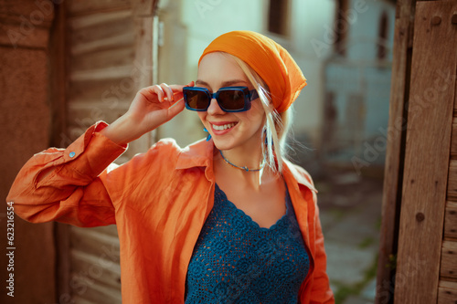 Fashionable happy smiling woman wearing trendy blue rectangular sunglasses, orange linen shirt, summer crochet top, posing in street of city. Copy, empty space for text