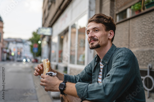 One man young adult modern caucasian male in the city in sunny day stand and eat sandwich fast food concept urban life copy space tourist eating in front of building wall real person