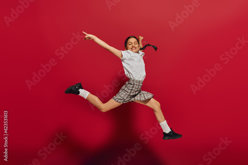 Full length of pleased and stylish preteen girl with hairstyle wearing t-shirt and plaid skirt while jumping and having fun on red background, hairstyle and trendy accessories concept