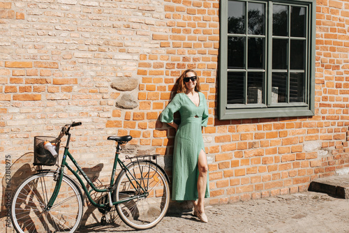 Kobieta w zielonej sukience oraz z zielonym rowerem stoi pod ceglaną ścianą w Gdańsku