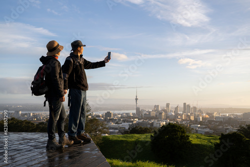 Couple taking photo with smartphone on Mt Eden summit at sunrise. Skytower in the background. Selective focus on people in foreground. Auckland.