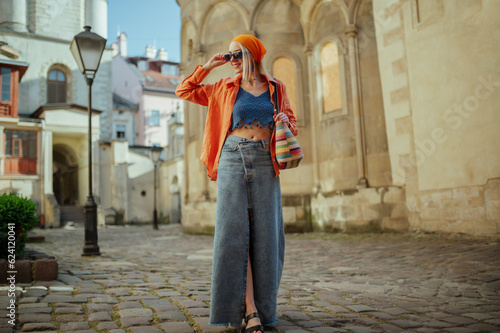 Fashionable happy smiling woman wearing trendy sunglasses, orange shirt, bandana, crop top, low waist denim maxi skirt, with wicker bag, posing in street of European city. Copy, empty space for text