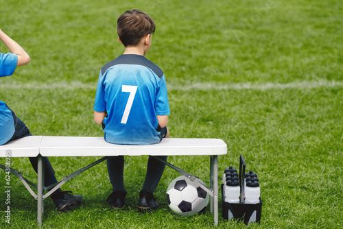 Schoolboy sitting on soccer bench. Young boy sitting on the substitute bench. Football sports competition game for children. The little boy in a blue jersey with the number seven