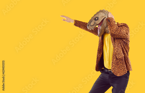 Man in funny reptile costume dancing on copy space background. Side view of happy young guy in leopard jacket and Halloween dinosaur mask dancing isolated on yellow copyspace background. Party concept