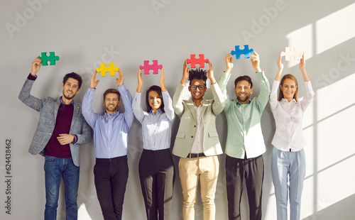 Group of young happy business people holding colorful puzzle parts. Coworkers, colleagues or team of staff standing in a raw together with jigsaw pieces with hands up on a gray wall background.