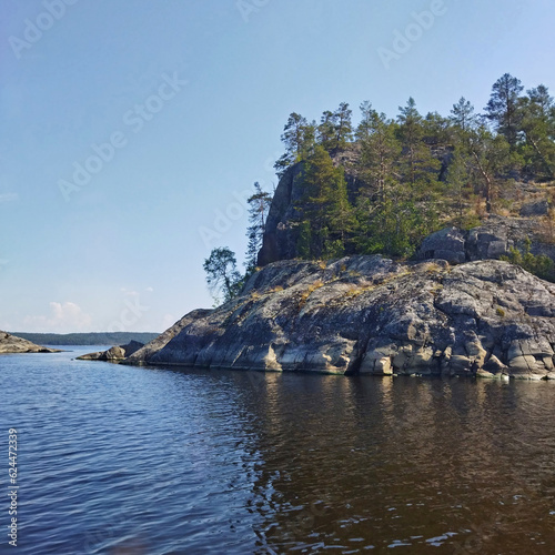 Beautiful summer nature of northern lake. Landscape with rocky coast, green trees, blue sky and reflection in calm water. Ladoga lake, Karelia, Russia.