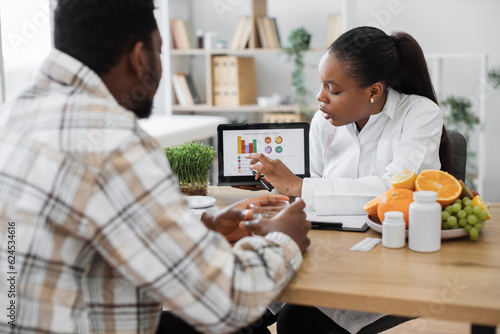 Over-shoulder view of african american woman analyzing patient's nutrition data on digital tablet in consulting room. Efficient dietitian assessing health needs of male client in medical centre.