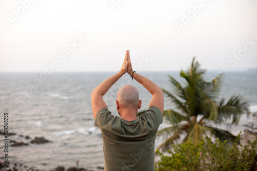 Rear view of vacationer folding palms together and lifting arms up, practicing yoga against seascape in front of sea.
