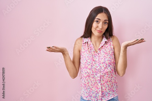 Young hispanic woman with long hair standing over pink background clueless and confused expression with arms and hands raised. doubt concept.