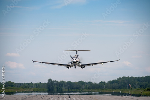 A small business turboprop Pilatus PC-12 on final with the landing gear starting to extend. Blue sky with haze and clouds in the background.