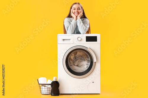 Pretty young woman leaning on washing machine on yellow background