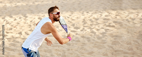 Dynamic image of young man playing beach tennis, hitting ball with racket. Outdoor training on warm summer day. Top view. Concept of sport, leisure time, active lifestyle, hobby, game, summertime, ad