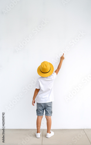 An child in summer clothes stands in front to the white wall, his hand raised and pointing a point on the wall