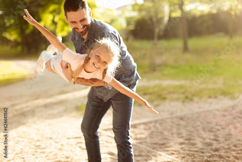 Family day outdoors. Happy middle aged father holding fearless daughter on hands, girl pretend flying outdoor in park