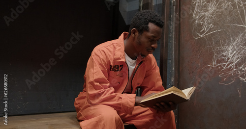 Upset african american prisoner in jail cell sits reads book looks at camera.