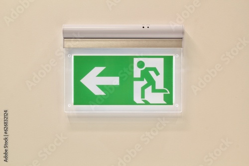 Close-up of green emergency exit sign showing the way to escape on wall. Fire exit in corridor of building. Fire prevention and house safety concept. Arrow pointing exit