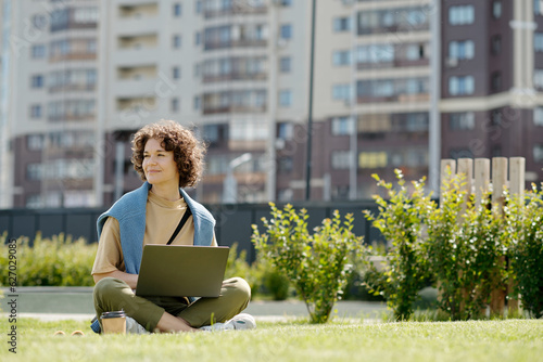 Young smiling woman with laptop sitting on green grass in park on sunny summer day and looking aside while enjoying rest and watching video