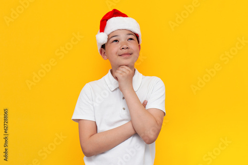 pensive little asian boy in santa hat imagines and dreams on yellow isolated background, korean child