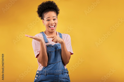 Portrait, space and a black woman pointing to her palm for the promotion of a product on a yellow background in studio. Smile, advertising or marketing with a happy young female brand ambassador