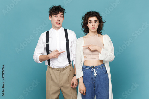 Portrait of scared young couple or friends standing and pointing at each other with index fingers, having frighten facial expression. Indoor studio shot isolated on blue background.