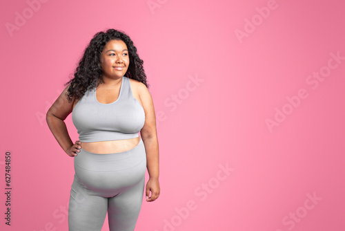 Young black plus size woman in sportswear standing over pink studio background, looking aside at free space and smiling