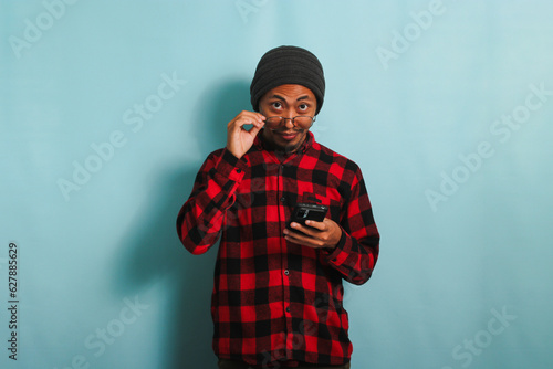 Young Asian man with a beanie hat and a red plaid flannel shirt is staring through his eyeglasses while holding a phone, isolated on a blue background