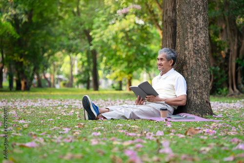 portrait happy senior man sitting against the tree and reading a book in the nature park, concept of senior activities,hobby,relaxing,health benefits in nature