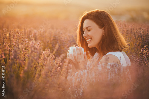 Young happy woman drinking herbal tea, sitting in a beautiful lavender field at sunset.
