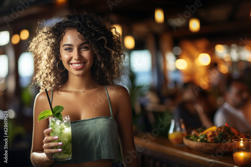 An attractive young woman holds a refreshing drink with a mint leaf on top, mojito, she is in a tropical restaurant near the beach, copy space