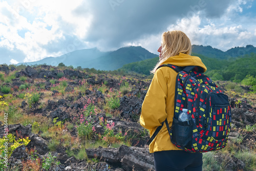 Hiking on tallest volcano in Continental Europe - Etna. Young woman in yellow raincoat standing on the mountain slope in front of panoramic view of Mount Etna.